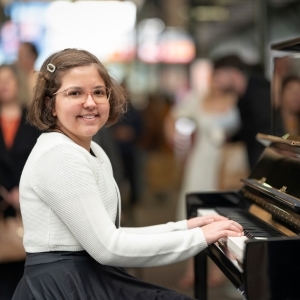 A young girl sitting in front of a piano with her hands on the keys looking at the camera and smiling.