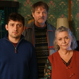 Promotional image for the show Can You Keep A Secret featuring three main characters—a younger man, an older man, and an older woman—standing together in a living room with floral wallpaper.