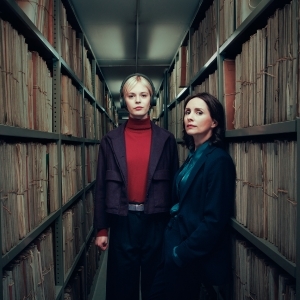 Two women stand in a narrow archive aisle flanked by tall shelves filled with old files; one woman wears headphones.
