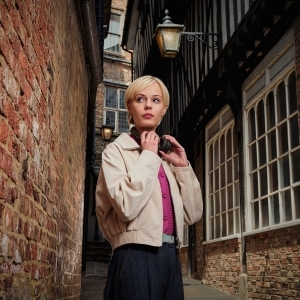 A young woman with short blonde hair stands in a narrow, historic brick alleyway, adjusting a pair of headphones around her neck.