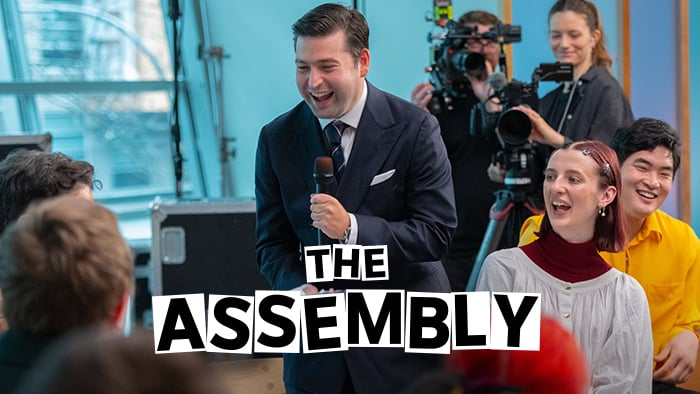 A man in a navy suit laughs into a microphone, surrounded by a smiling audience and a camera crew in a studio. The words THE ASSEMBLY are centered in large, bold block letters at the bottom.