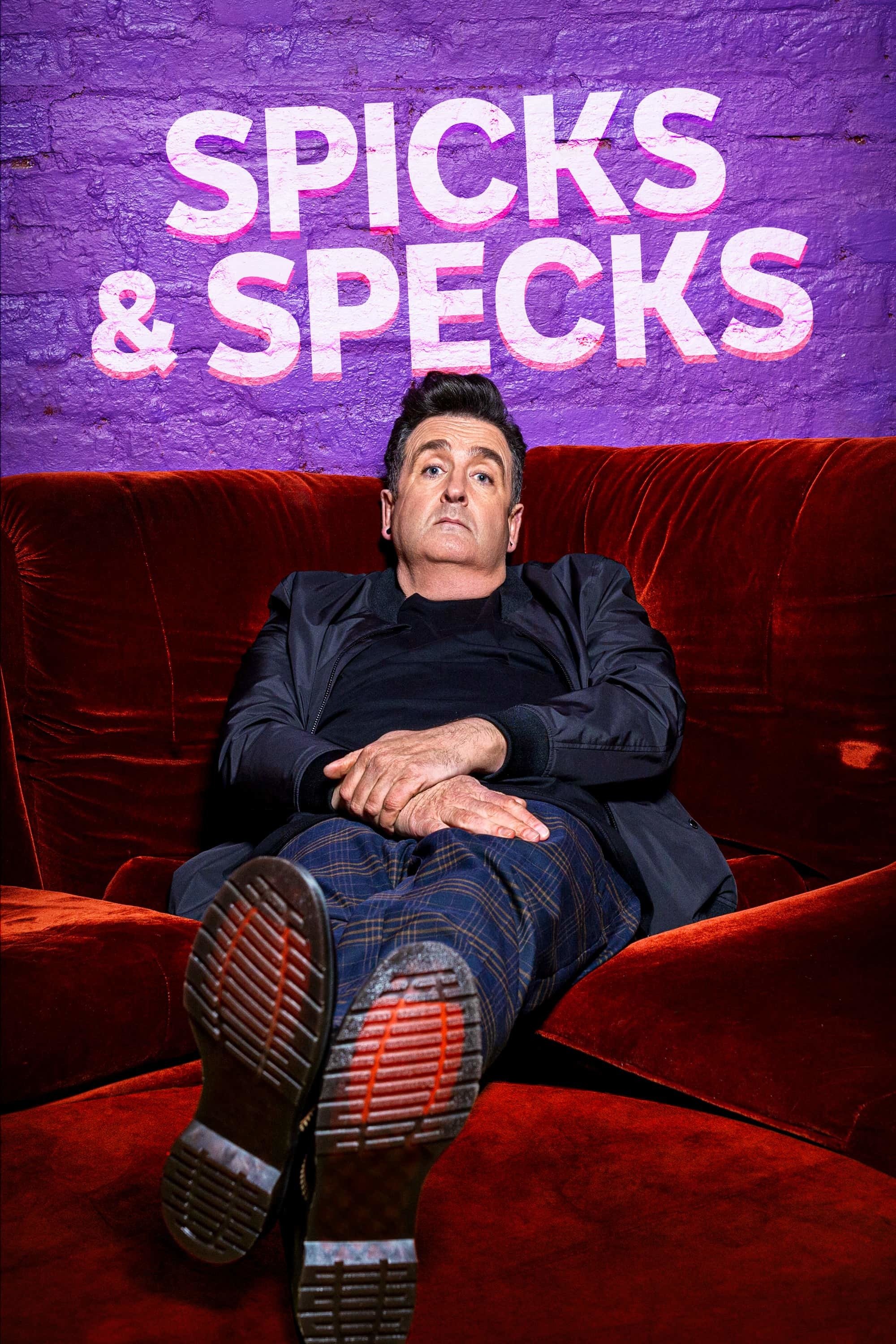 Host Adam Hills leans back on a red couch, feet toward the camera, under a neon-style Spicks and Specks sign.