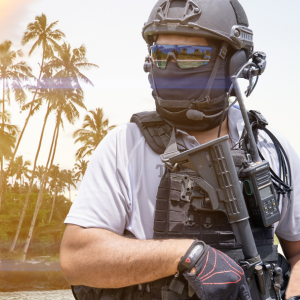 A tactical officer in full gear, including a helmet, face mask, and sunglasses, stands before a tropical backdrop of palm trees.