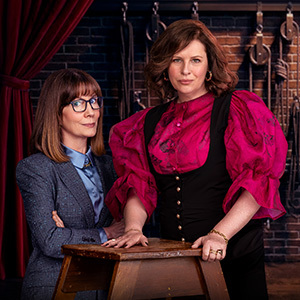 Banner for ABC iview streaming service with download button and show image for Bad Company showing Kitty Flanagan in a grey patterned blazer and glasses with arms crossed, beside her is Anne Edmonds wearing a vibrant magenta puff-sleeve blouse and black vest leaning on a wooden stool.