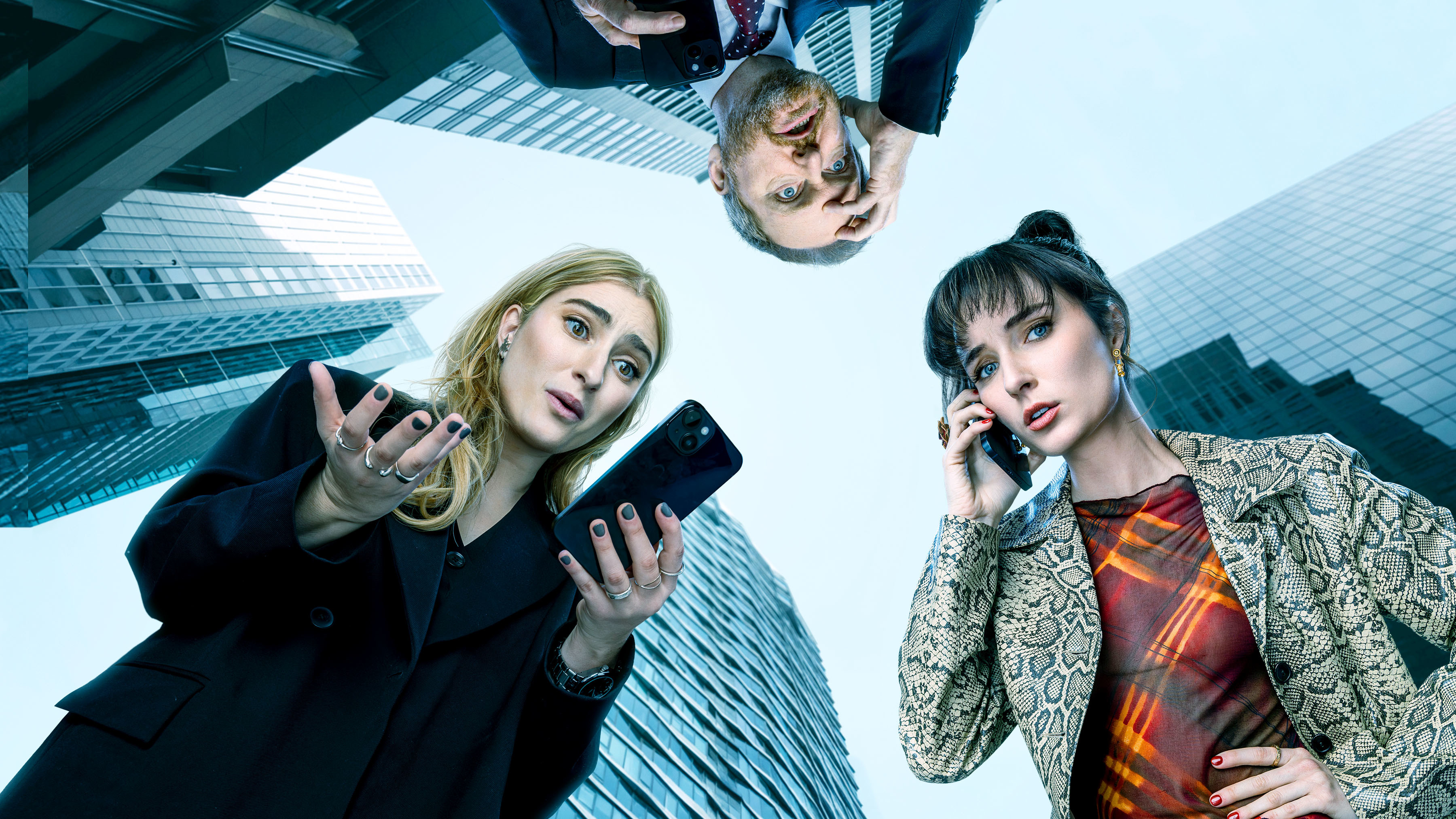 A low-angle shot looking up at three people against a backdrop of towering city skyscrapers. A young woman with blonde hair in a black blazer holds a phone outward with a surprised expression, a young woman with dark hair in a snakeskin jacket holds a phone to her ear, and a man in a suit appears upside down at the top of the frame.