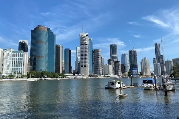 A view of an Australian city skyline along a calm waterfront, featuring tall skyscrapers in varying architectural styles and small boats near the edge of the water.